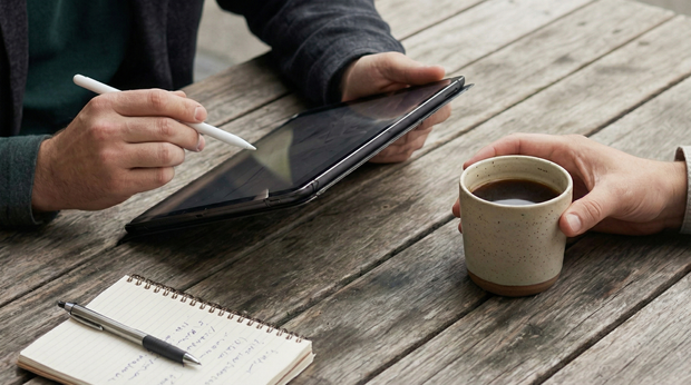 Two people at a wooden table with a tablet, notebook, pen, and coffee cup.